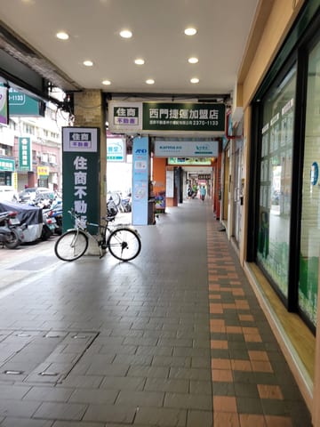 A bicycle parked beneath a long covered sidewalk lined with shops in Taipei