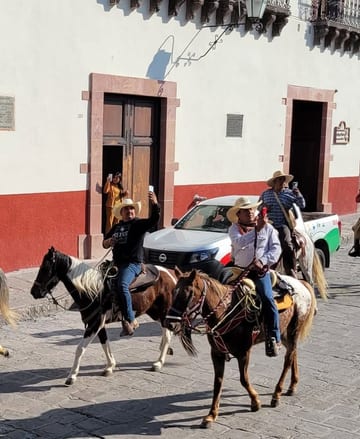 Cowboys riding horses along a cobblestone street past white and red buildings